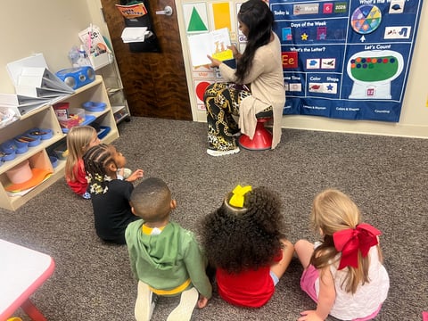 Teacher watering plant with four young children sitting on classroom floor watching, educational bulletin board in background