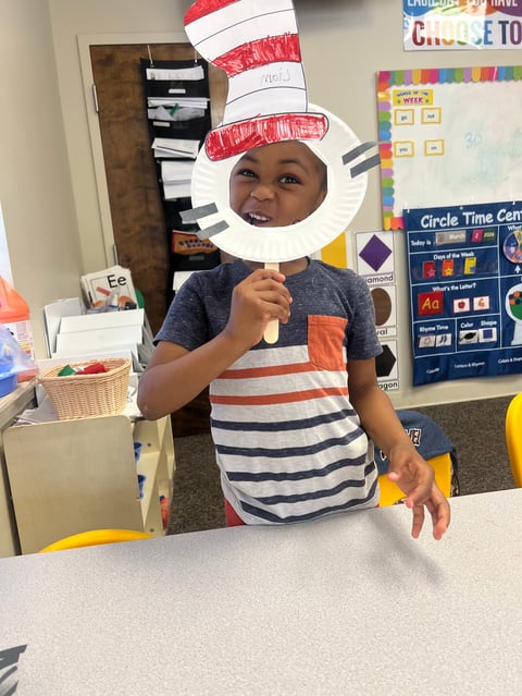 Young child in classroom wearing striped shirt and paper Cat in the Hat costume with tall red and white hat