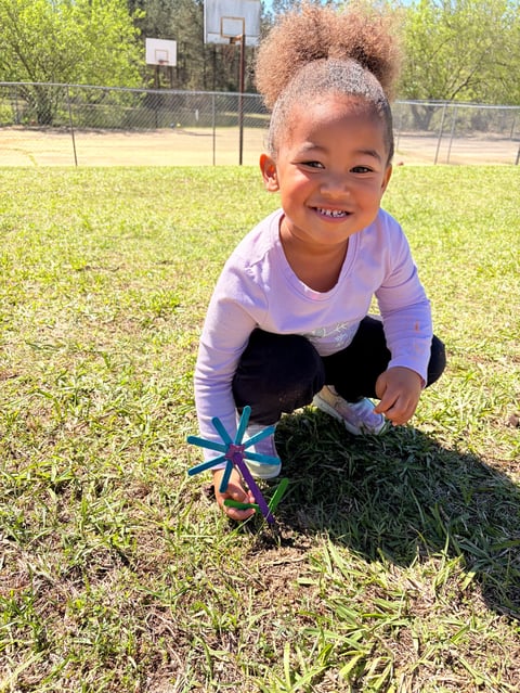 Young child smiling at outdoor basketball court while playing with blue toy windmill in grass