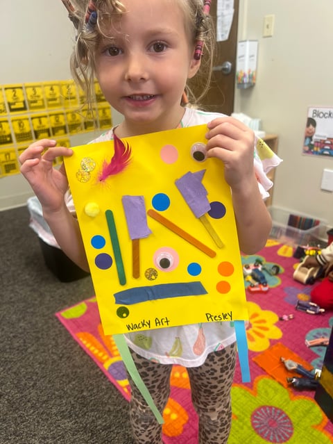 Young child displaying colorful wacky art face craft made with yellow paper, foam shapes, and googly eyes