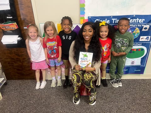 Adult woman posing with six young children in a classroom, holding a children's book