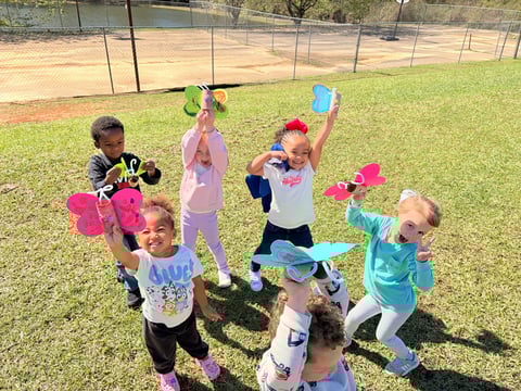 Group of young children in colorful outfits playing with foam toys on a grassy field near a baseball diamond