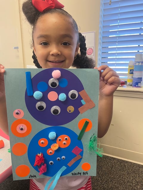 Young girl with red bow smiling while holding colorful handmade craft project with painted circles and googly eyes