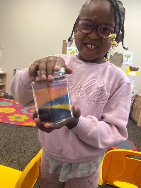 Young child wearing purple glasses and pink sweater holding up a colorful layered sand art jar in a classroom setting