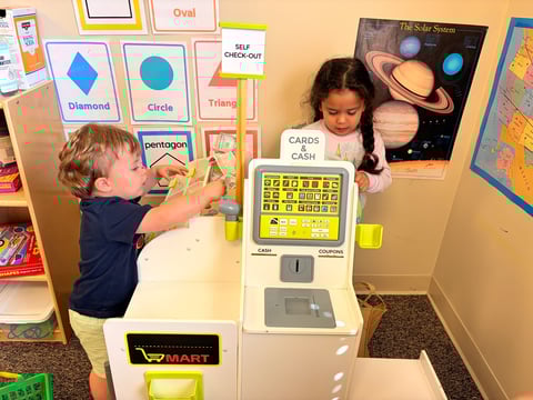 Two young children interact with educational displays in a classroom, including a toy cash register and learning posters with shapes and planets on the walls