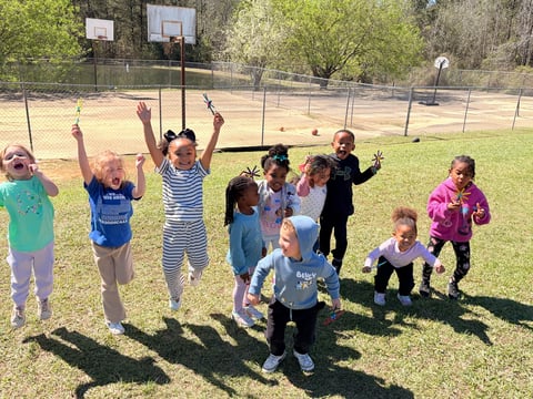 Group of children playing together in a sunny park with basketball hoops and a fenced area in the background