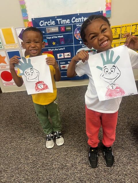 Two young children smiling and holding handprint art projects in a classroom with Circle Time Center board behind them