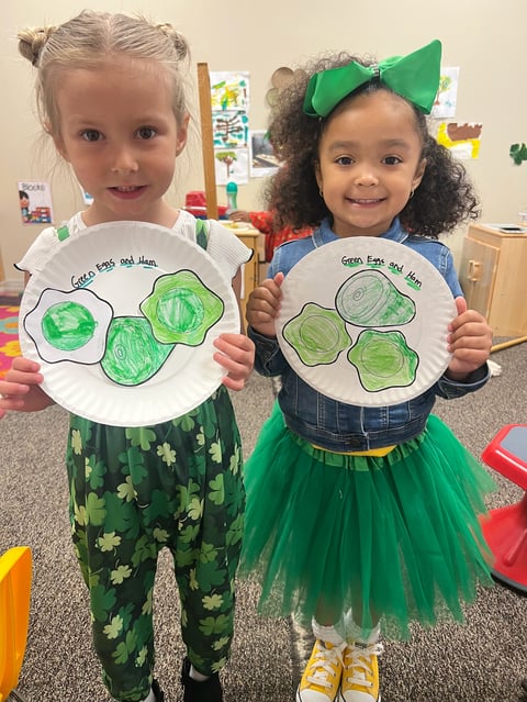 Two young children in classroom holding paper plate crafts decorated with green painted vegetables, both smiling at camera