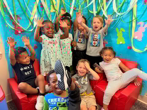 Group of young children celebrating at a colorful birthday party with streamers and balloons, sitting and standing on red cushions against a bright blue backdrop