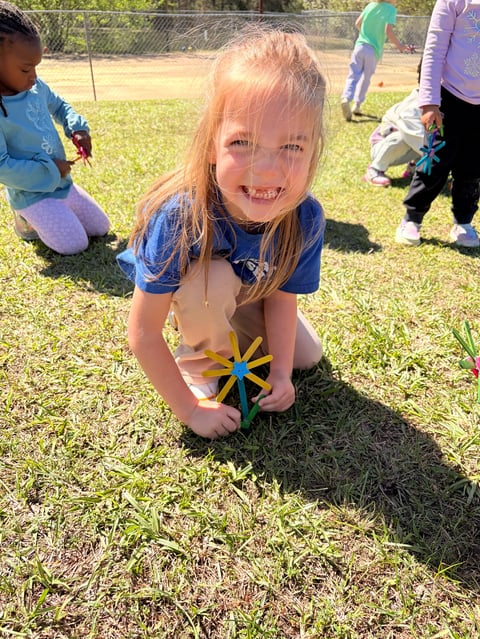 Young child with red hair smiling at camera while holding a colorful pinwheel toy in a grass field with other children and adults in background