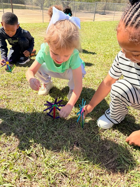 Children planting a small tree together in a grassy outdoor area, with a chain-link fence visible in the background