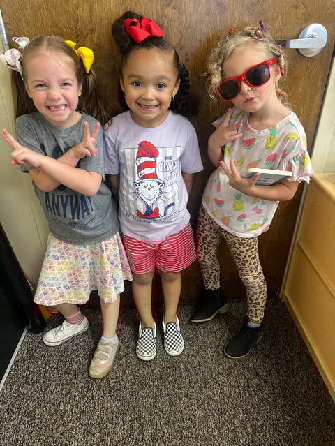 Three young girls posing together indoors wearing colorful outfits and hair accessories, with one wearing sunglasses