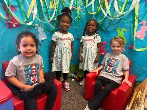 Four young children sitting on red chairs in front of a colorful turquoise wall decorated with ribbons and paper flowers